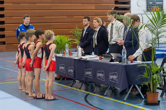 Young male gymnasts in red-black uniforms stand at attention before Gymfed judges at an official scoring table indoors.