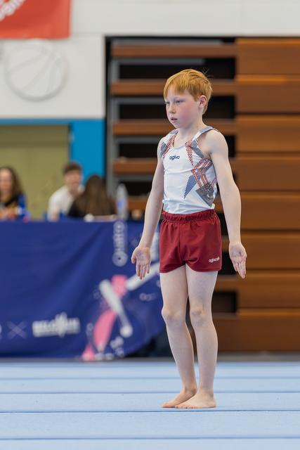 Young red-haired boy stands barefoot on the floor mat, focused and composed, arms slightly extended, ready to perform.