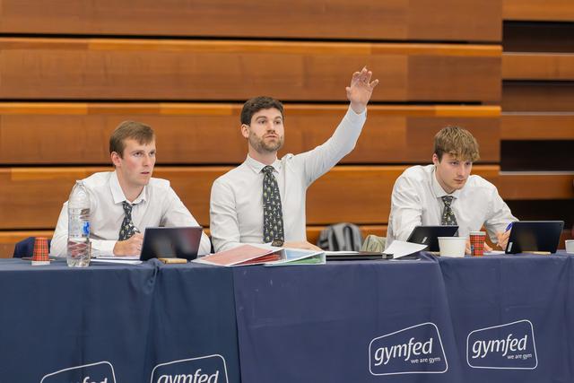 Three male judges at a gymfed table, center judge raises hand confidently while others review notes on laptops.