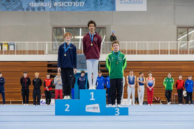 Three young gymnasts stand proudly on a Gymfed podium, gold medalist centered, all wearing medals in a Belgian sports hall.