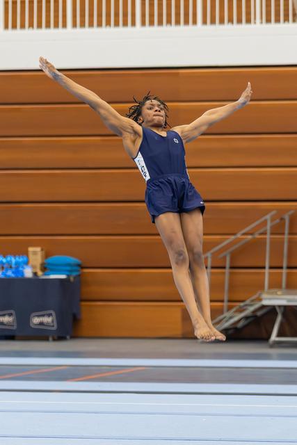 Young gymnast leaps mid-air with arms outstretched and focused expression during floor exercise in indoor gym.