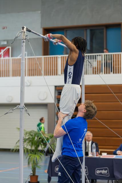 Coach lifts gymnast to reach the high bar in a busy indoor gymnastics hall, focused and determined.