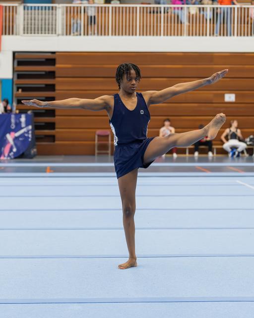 Young male gymnast holds a precise arabesque on one leg, arms extended, focused expression on a blue competition floor.