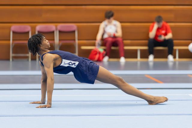 Young male gymnast in blue leotard performs a split stretch on floor mat, focused and controlled, indoors.