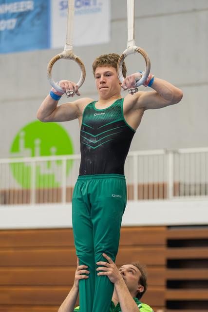 Young male gymnast grips still rings with intense focus, supported from below by a coach in a gym.