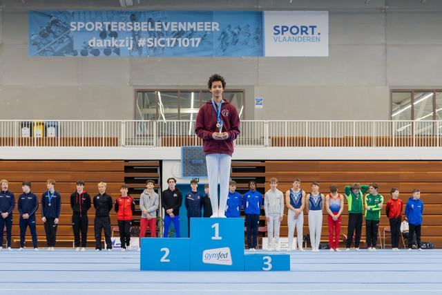 Young gymnast stands proudly on the first-place podium, medal around neck, at a Gymfed indoor gymnastics competition in Belgium.