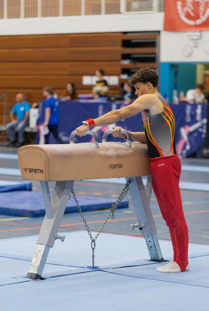 Young male gymnast grips pommel horse handles, focused and composed, preparing his routine in a busy gymnasium.