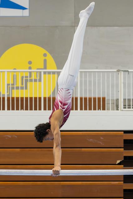 Young gymnast performs a perfect handstand on the balance beam, legs fully extended upward, in an indoor sports hall.