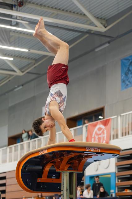 Male gymnast holds a precise handstand on a Spieth vault, legs extended upward, in a packed indoor gymnasium.