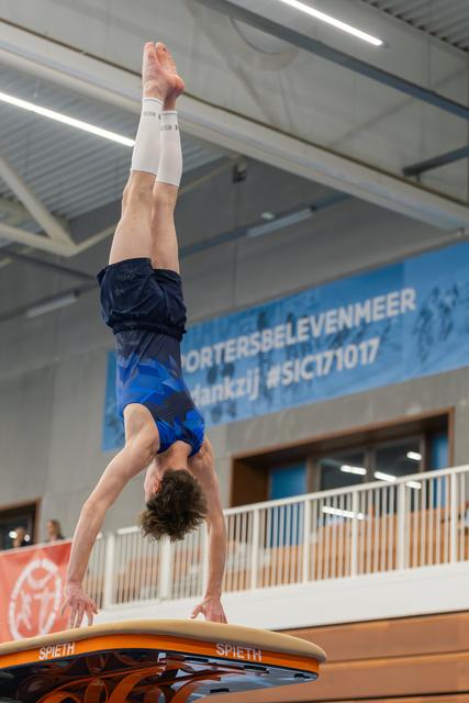 Young male gymnast holds a perfect handstand on a Spieth vault table, legs fully extended, in an indoor sports hall.