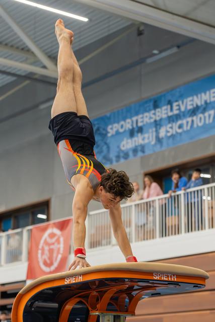 Young male gymnast performs a precise handstand on a Spieth vault, legs perfectly vertical, in an indoor sports hall.
