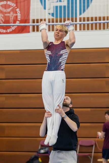 Young male gymnast grips the high bar with focused determination while his bearded coach spots him from below in a gym.