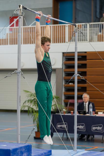Young male gymnast hangs from the high bar with intense focus, wearing green trousers and a dark leotard at a Gymfed event.