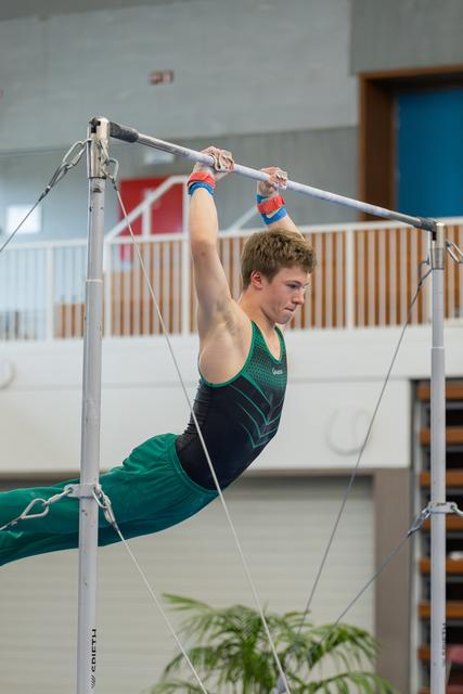 Young male gymnast hangs in a pike position on the high bar, face set with fierce concentration during indoor competition.