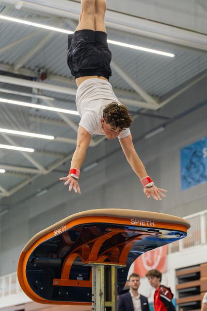 Young male gymnast mid-flight above a Spieth vault, body inverted, arms reaching downward with intense focus.