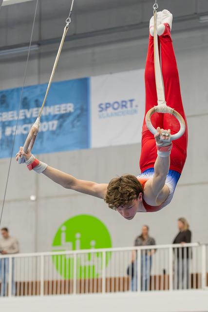 Young male gymnast performs an inverted iron cross on still rings, arms wide, body arched, at an indoor gymnastics meet.