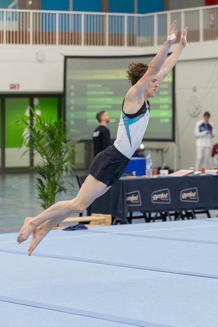 Young male gymnast soars above the floor mat, arms stretched high, body arched in a powerful dramatic leap.