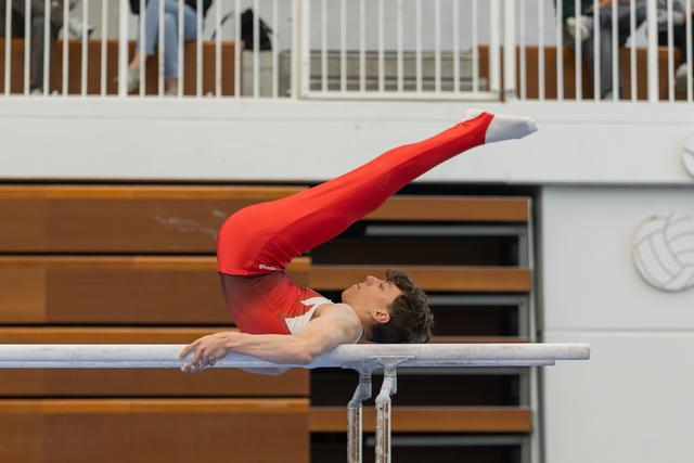 Male gymnast in red leotard performs a dramatic back arch on parallel bars, legs extended skyward, focused expression.