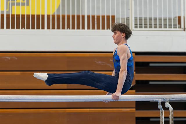 Young male gymnast holds an L-sit on parallel bars, legs extended forward, focused expression in a gymnasium.