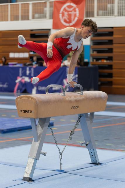 Young male gymnast in red suit performs dynamic move on pommel horse, legs extended mid-air with focused expression.
