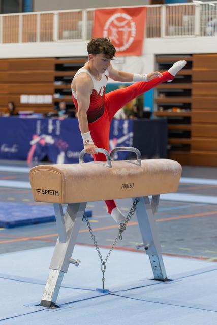 Young male gymnast performs on pommel horse, leg raised high, focused expression, indoor gymnastics hall.