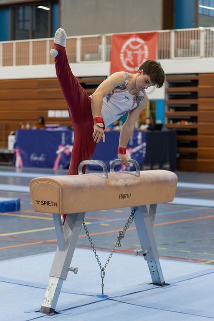 Young male gymnast performs on pommel horse, leg raised high, concentrating intensely in an indoor gymnasium.