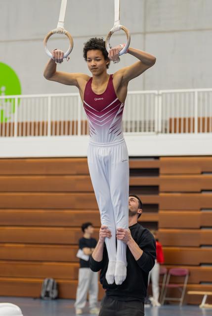 Young male gymnast grips still rings with focused expression while coach below steadies his legs in gym hall.