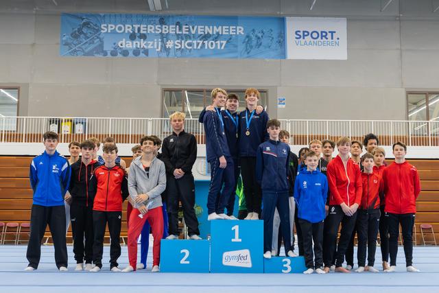 Young male gymnasts celebrate on a 1-2-3 podium at a Gymfed competition, gold medalists embracing with joy.
