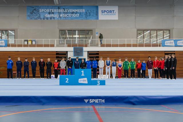 Young gymnasts line up beside a 1-2-3 podium inside a Sport Vlaanderen gymnastics hall, posing formally for awards.