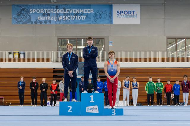 Three young gymnasts stand on a podium, 1st place looks proud, 2nd smiles, 3rd stands quietly at a Gymfed event.