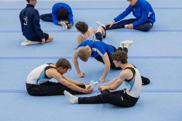 Young male gymnasts stretch and assist each other on a blue gymnasium floor, relaxed and focused during warm-up.
