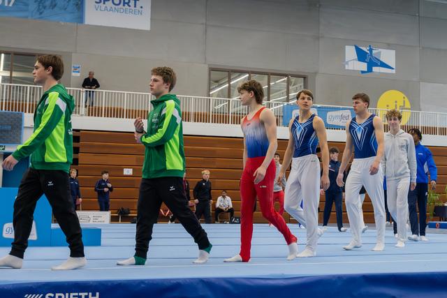 Male gymnasts in green and blue leotards walk across the floor mat during a gymnastics meet in Belgium.