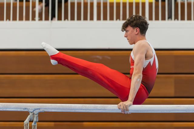 Young male gymnast performs on parallel bars, legs raised with intense focus, wearing red Canadian-style leotard in gymnasium.
