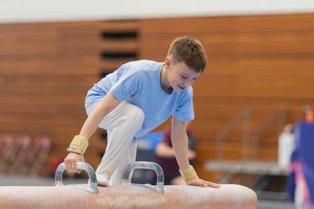 Young gymnast grips pommel horse handles with wrist guards, leaning forward with concentration during a gymnastics event.