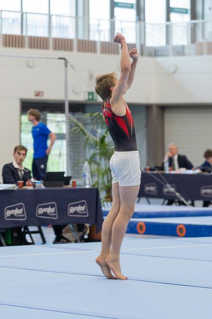 Young male gymnast stands tall on the floor, both fists raised overhead in a triumphant finishing pose at a Gymfed meet.