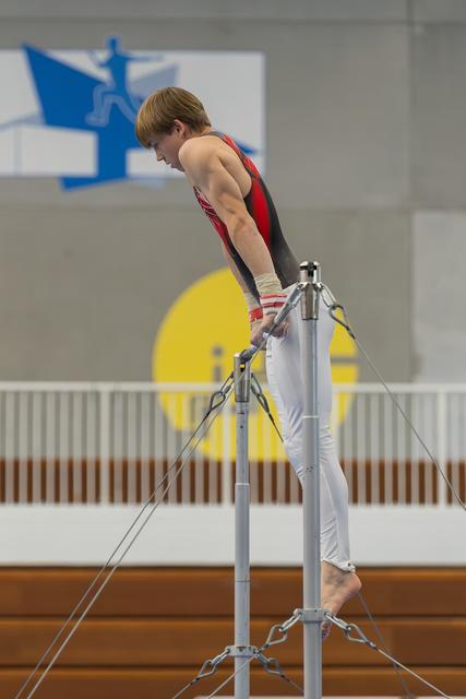 Young male gymnast stands atop the horizontal bar, leaning forward with intense focus in a gymnasium.