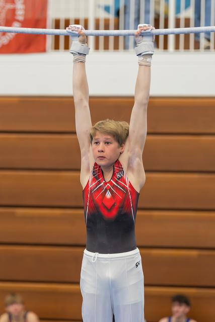 Young male gymnast hangs from the high bar with focused concentration, wearing a red and black leotard in a gymnasium.