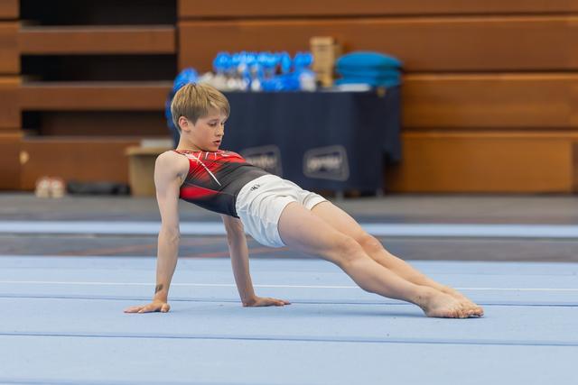 Young male gymnast performs a side plank hold on the floor exercise mat, focused and controlled.