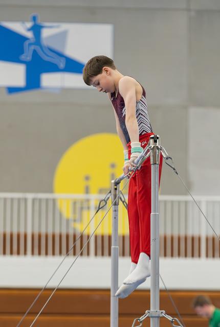 Young male gymnast grips the high bar with intense focus, body angled downward in a gymnastics hall.