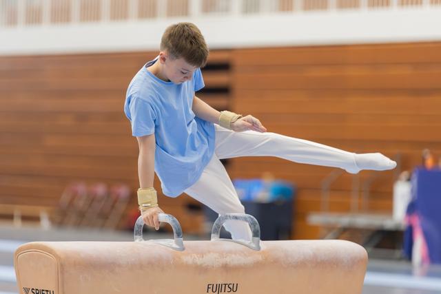Young boy in blue tee concentrates intensely while performing on a Fujitsu pommel horse, leg raised mid-routine.