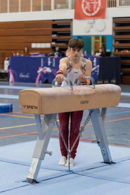 Young male gymnast grips the pommel horse handles with intense focus during an indoor gymnastics event.