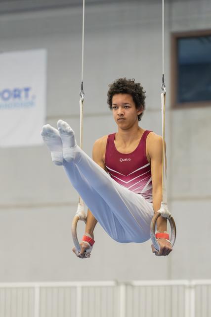 Young male gymnast holds a tuck position on still rings, wearing a red Quatro leotard, with intense concentration on his face.