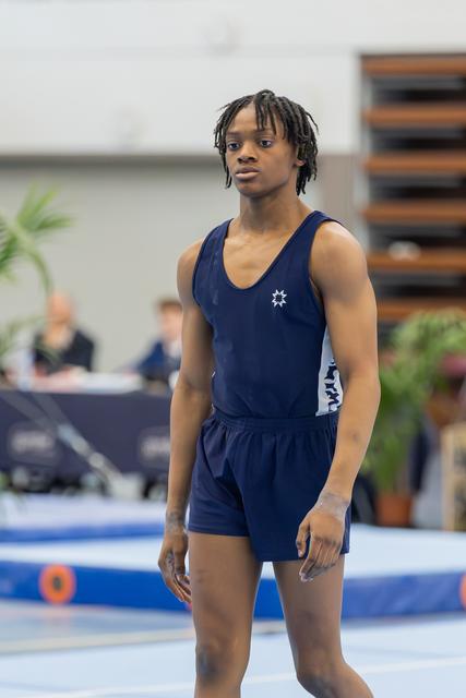 Young male gymnast in navy leotard stands composed and focused on the floor exercise mat, ready to perform.