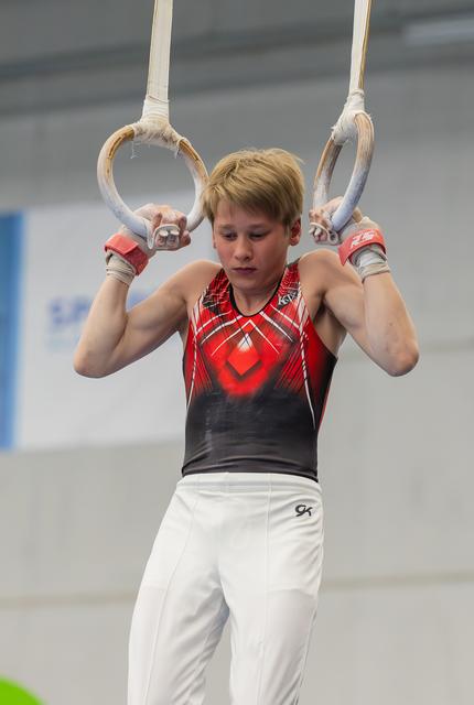 Young male gymnast holds steady on still rings, focused expression, wearing red and black leotard in indoor gym.
