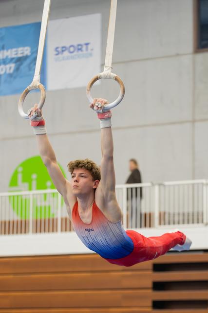 Young male gymnast hangs with intense focus on still rings, body extended horizontally in a dramatic hold.