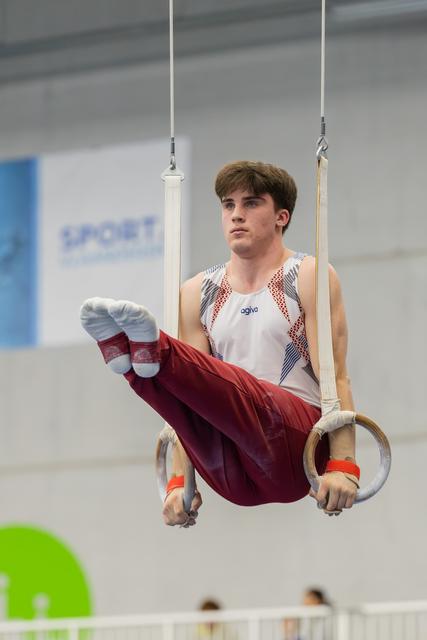 Young male gymnast holds a tuck position on still rings, focused expression, wearing agiva kit in a sports hall.
