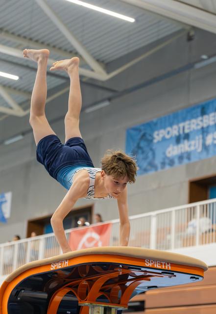 Young male gymnast performs a handstand on a Spieth vault, legs extended upward, focused expression, indoor sports hall.