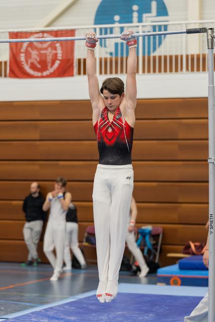 Young male gymnast hangs from the high bar with arms extended, wearing a red and black leotard, focused expression.
