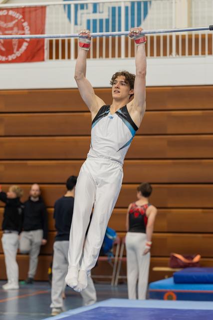 Young male gymnast hangs from the high bar with focused expression, wearing white and teal leotard in a gymnasium.