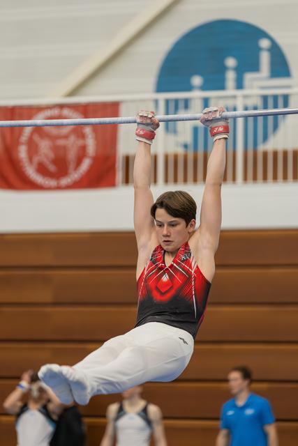 Focused teenage male gymnast hangs from the high bar, legs raised, wearing red and black leotard in a gymnasium.